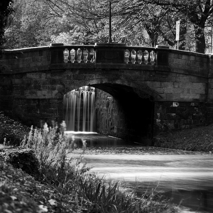 A stone bridge with a balustrade crosses over a river with a waterfall, surrounded by trees.