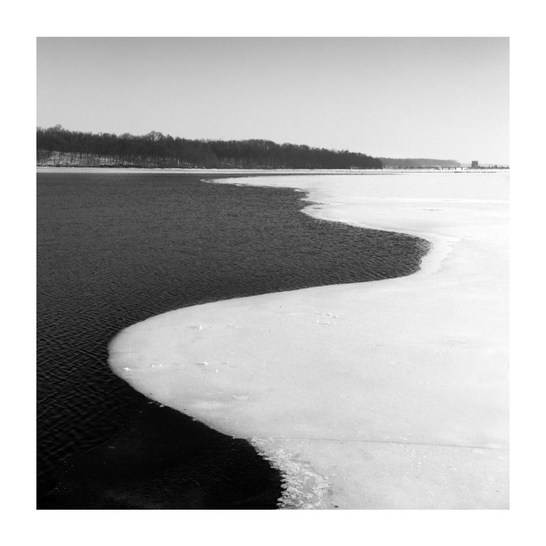 A body of water with a wavy line of ice and snow on one side and a distant treeline on the other.