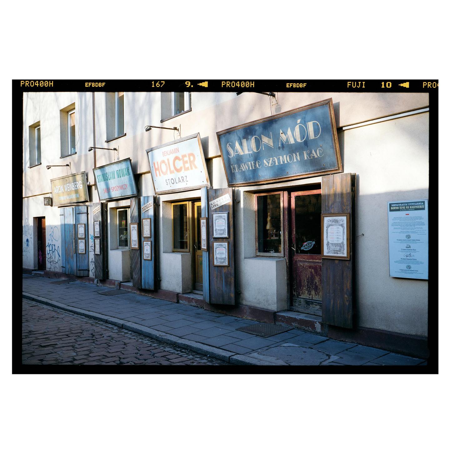 A row of storefronts with various signs and closed wooden shutters line a cobblestone street.