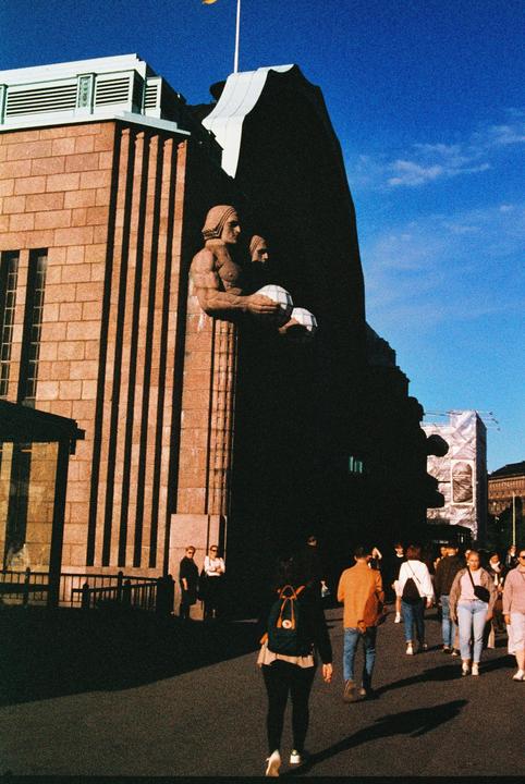 A crowd of people walk past a large building with two statues of men holding spheres.