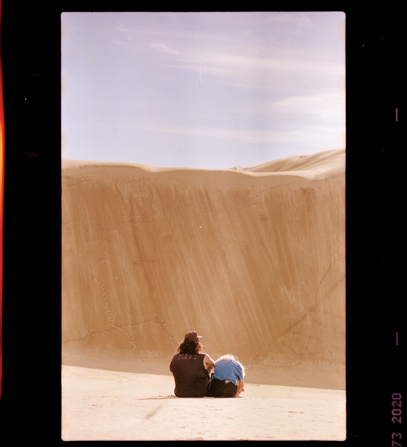 A couple sits on a dune looking at a large sand dune.