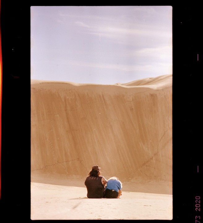 A couple sits on a dune looking at a large sand dune.