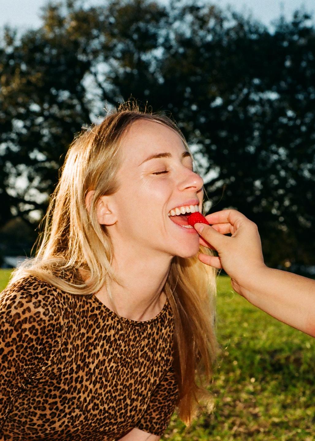 A woman with closed eyes and a wide smile is being fed a strawberry by another person.