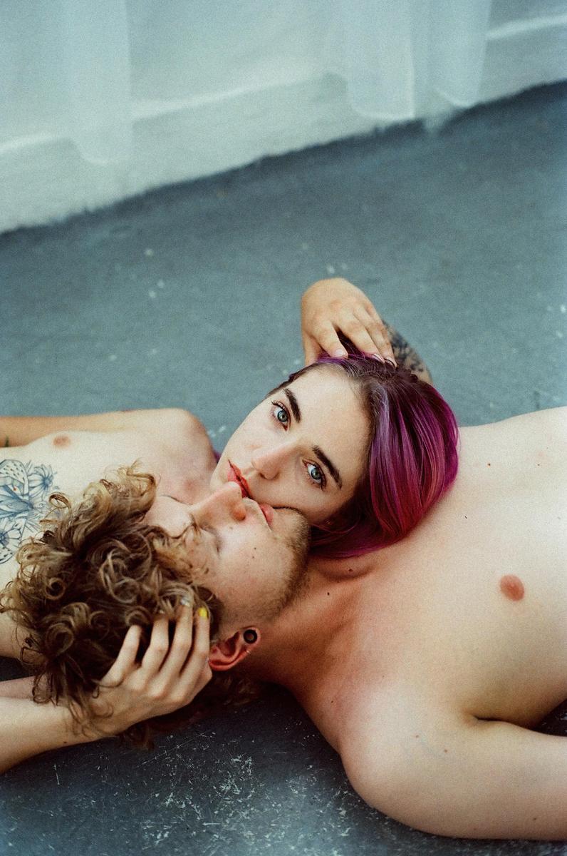 A couple lies on the floor, with the woman resting her head on the man's chest and looking at the viewer.