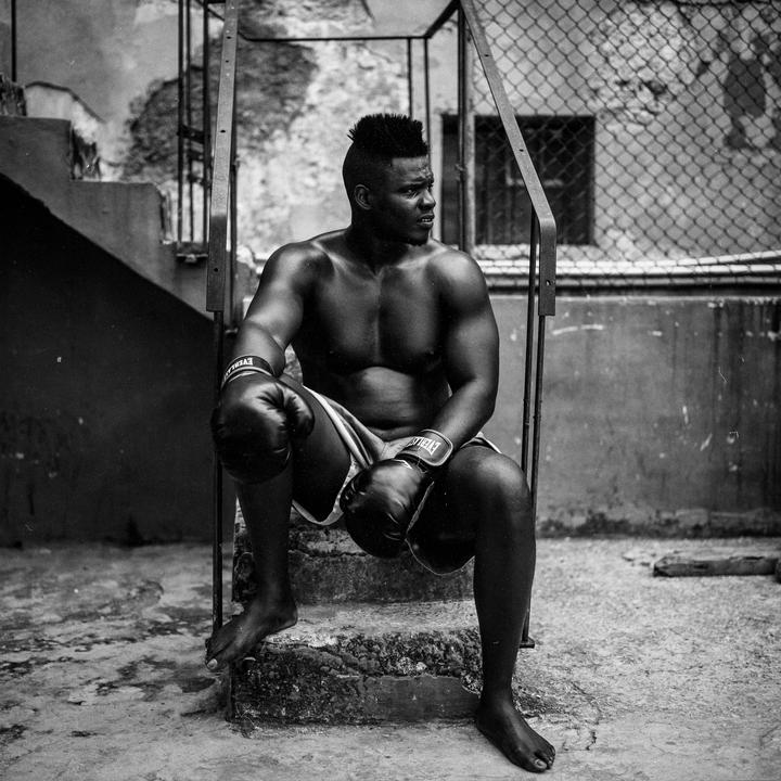 A man wearing boxing gloves and shorts sits barefoot on concrete steps next to a chain-link fence.