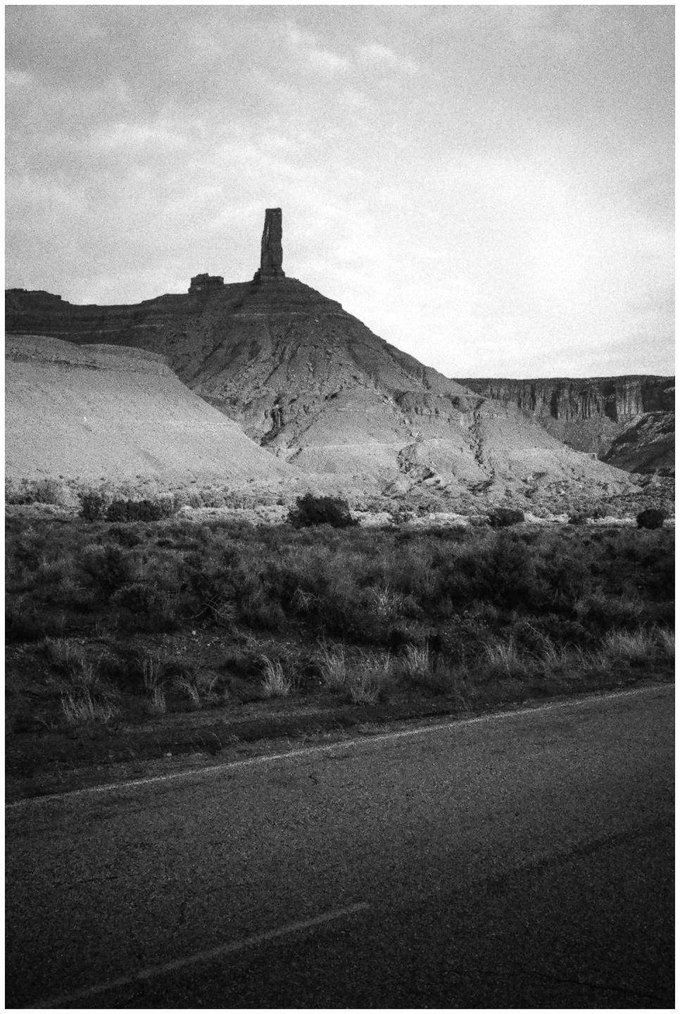 A road runs along the bottom of the frame, with a tall, thin rock formation on a hill in the background.