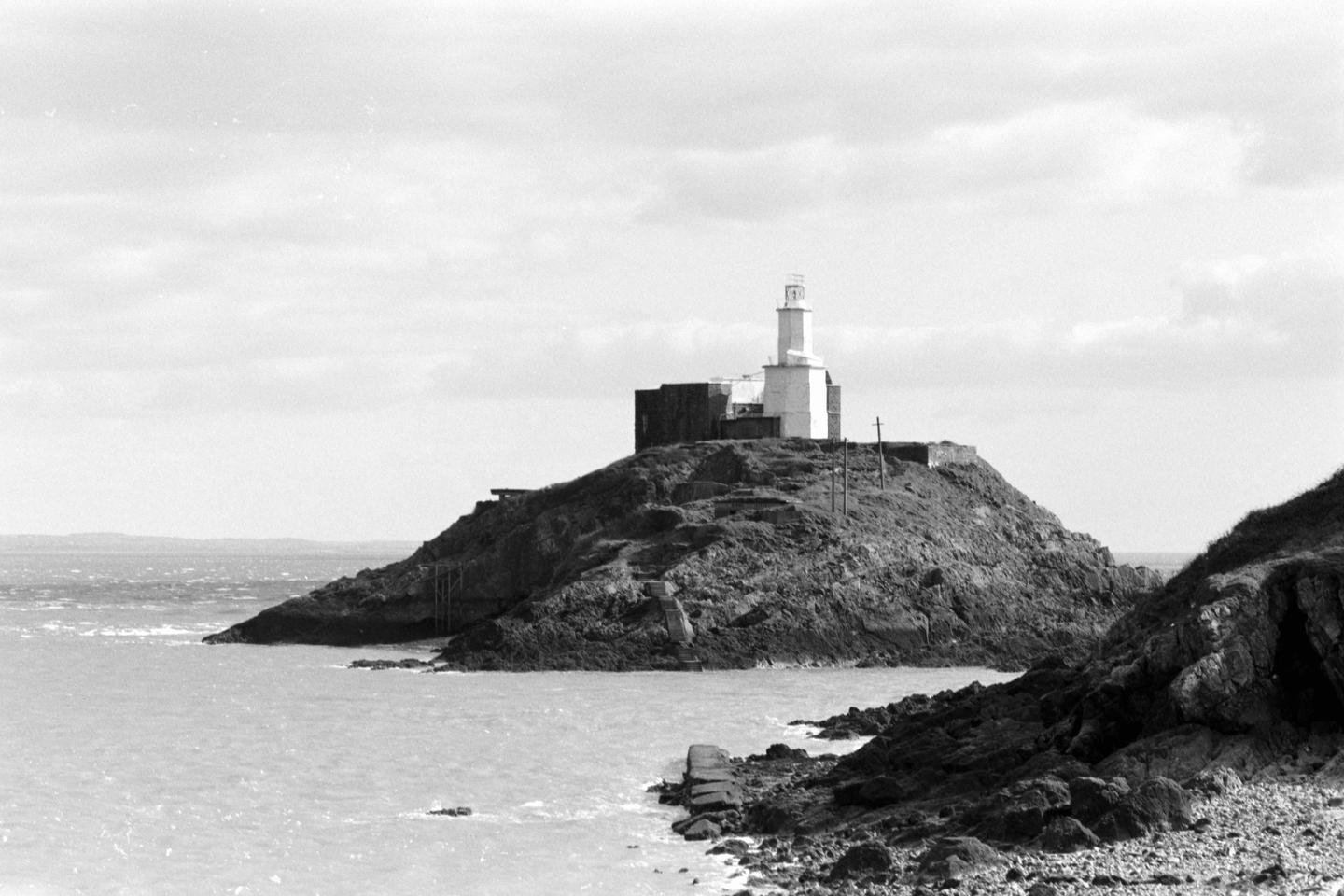 A white lighthouse and building stand on a rocky hill overlooking the sea under a cloudy sky.