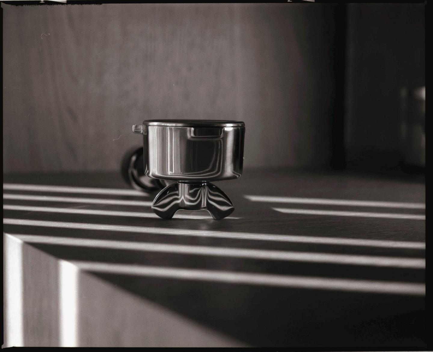 A metal espresso portafilter sits on a surface with strong shadows cast across it.