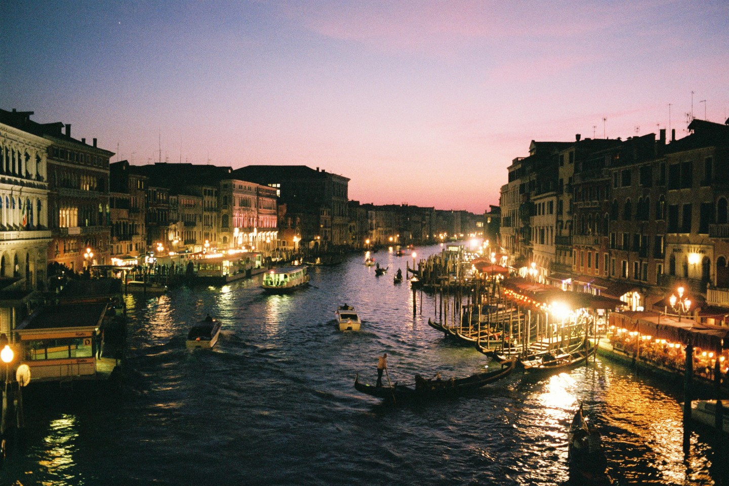 A canal in Venice is filled with boats and gondolas, with buildings lit up on both sides under a purple dusk sky.