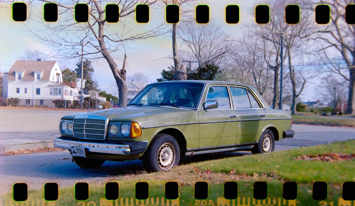 A green sedan is parked on a grassy verge next to a street with houses and bare trees in the background.