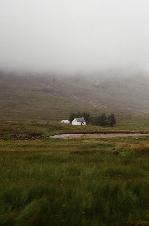 A small white house and shed sit in a green valley with a river and bridge, shrouded in fog.