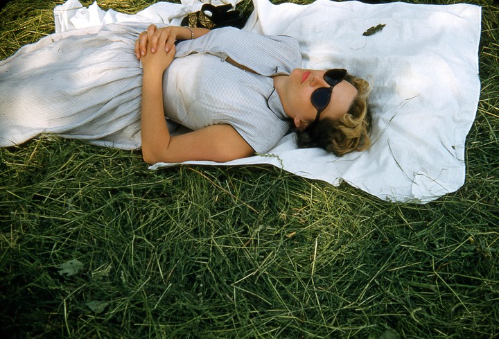 A woman wearing sunglasses and a striped dress is lying on a white blanket in the grass.