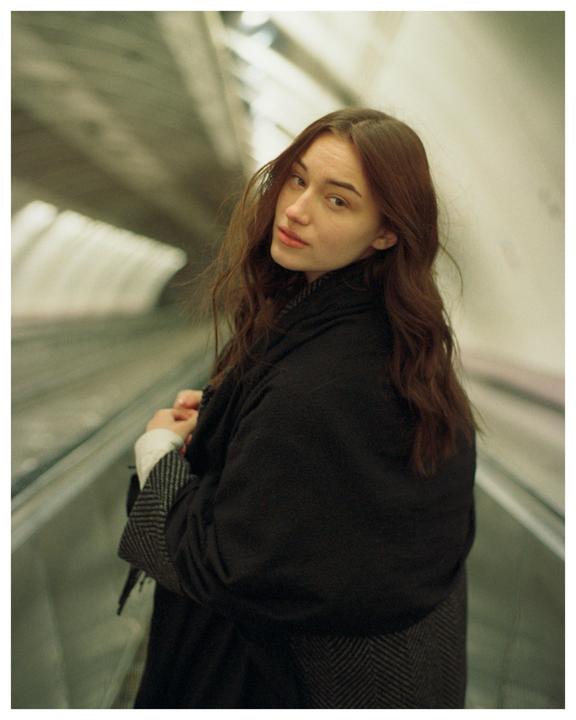 A woman with long brown hair looks over her shoulder while standing on an escalator.