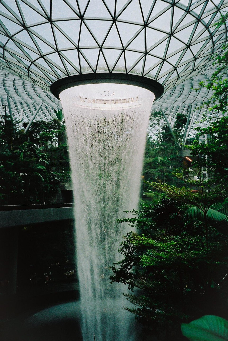 A large indoor waterfall cascades from a circular opening in a glass-paneled domed ceiling, surrounded by lush green foliage.