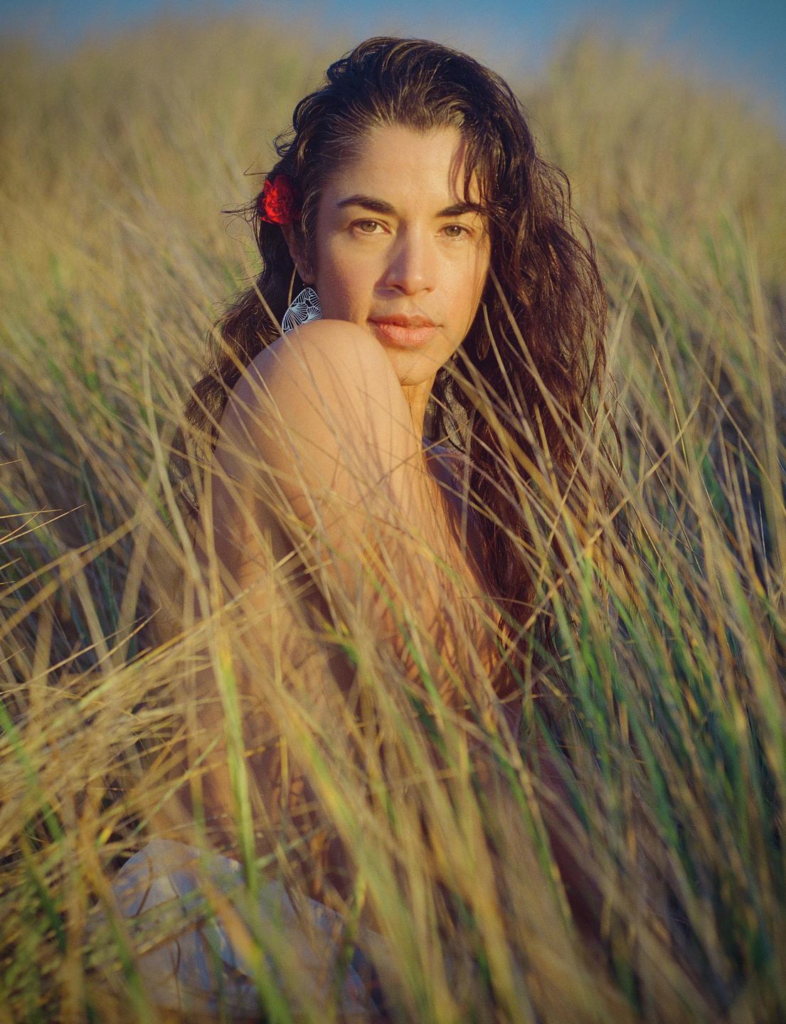 A woman with long hair and a red flower in her hair is sitting in tall grass.