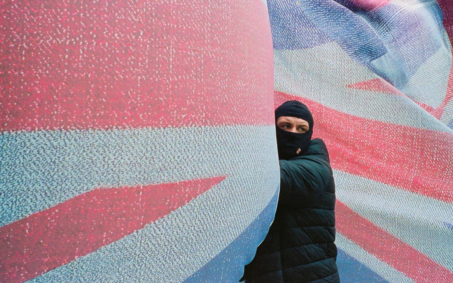 A man wearing a balaclava and a puffer jacket stands next to a large British flag.