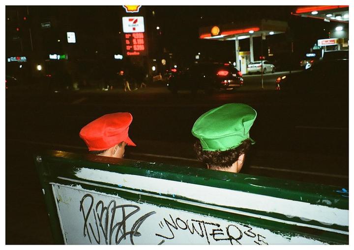 Two people wearing red and green hats are seen from behind, looking out at a street scene at night with a gas station and cars.