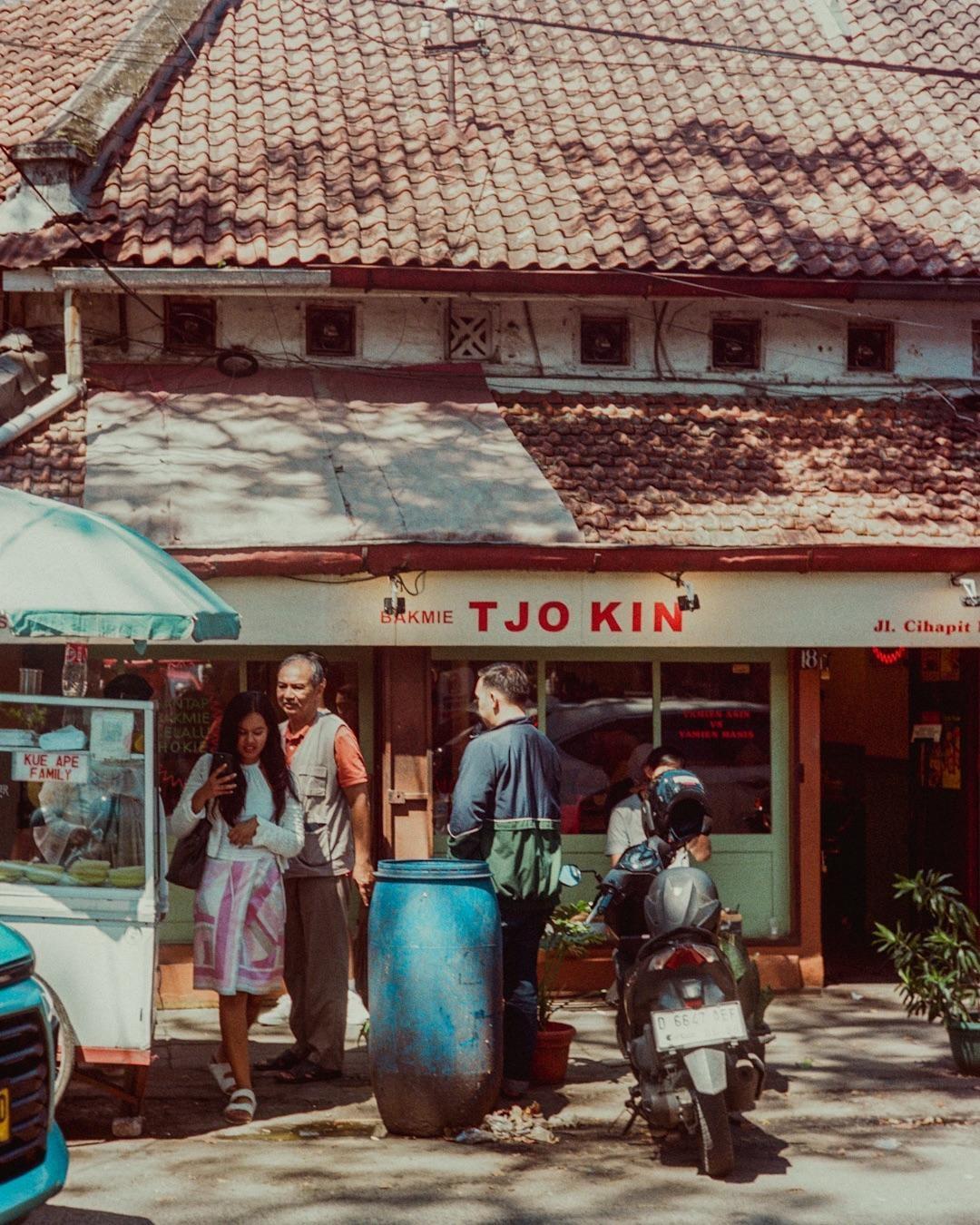 A group of people are gathered outside a storefront with a red tiled roof and a sign that reads "Bakmie Tjo Kin."