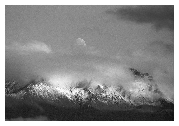 A moon is partially obscured by clouds above a snow-capped mountain range.