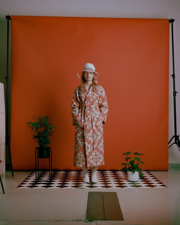 A woman in a patterned coat and white hat stands on a checkered floor in front of an orange backdrop, flanked by two potted plants.