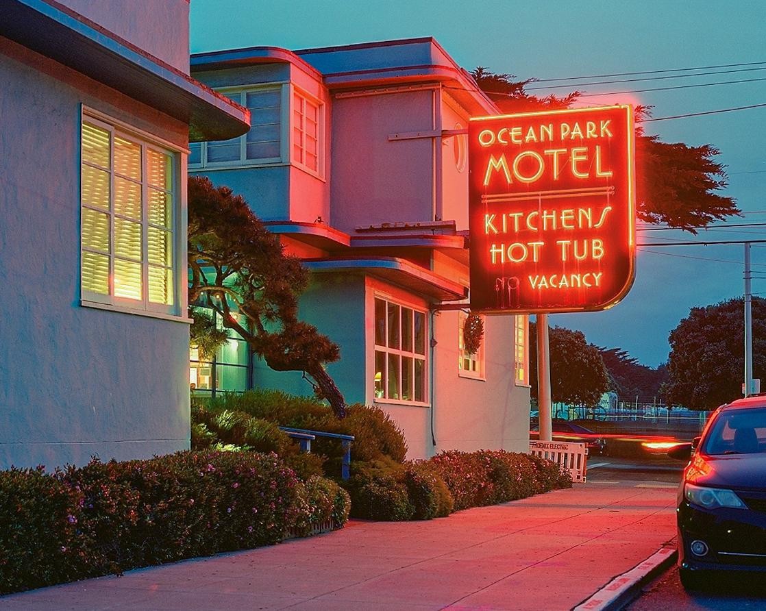 A neon sign for the Ocean Park Motel is illuminated at dusk, casting a red glow on the surrounding buildings and sidewalk.
