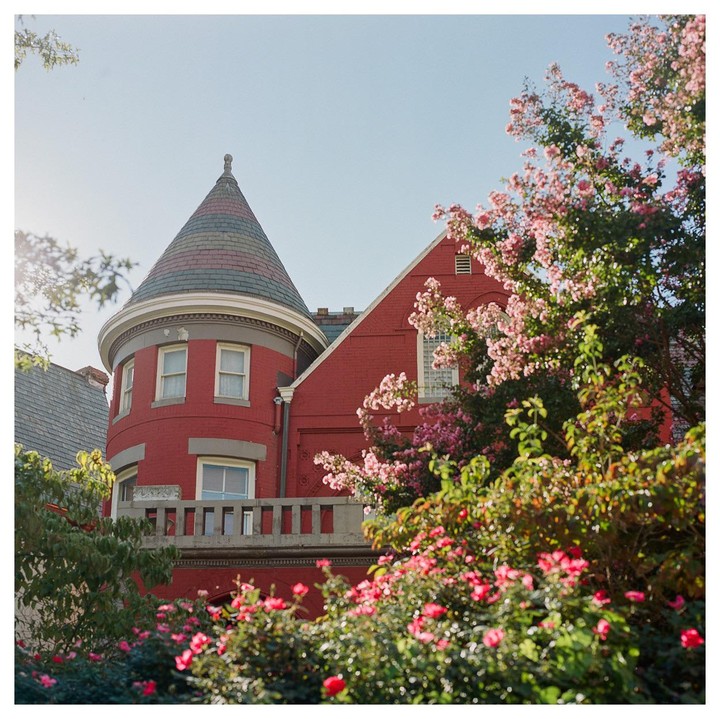 A red brick building with a conical roof is surrounded by flowering bushes and trees.