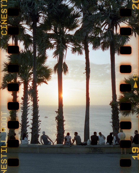 A group of people sit on a wall watching the sunset over the ocean, framed by palm trees.