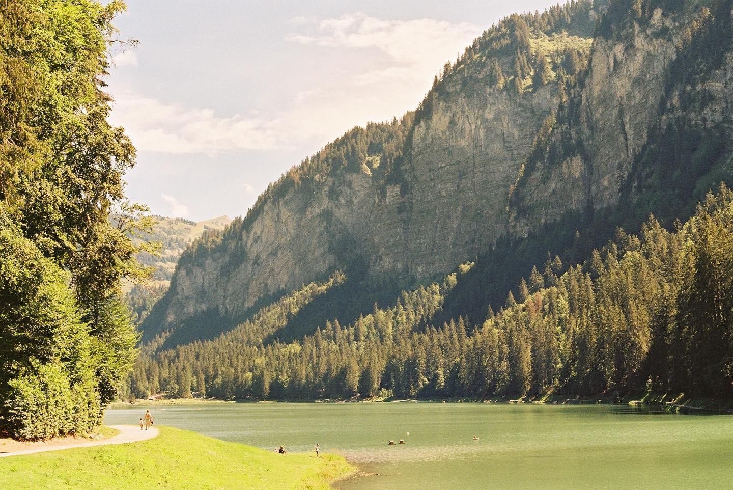 A woman and child walk along a path next to a lake surrounded by a forest and mountains.