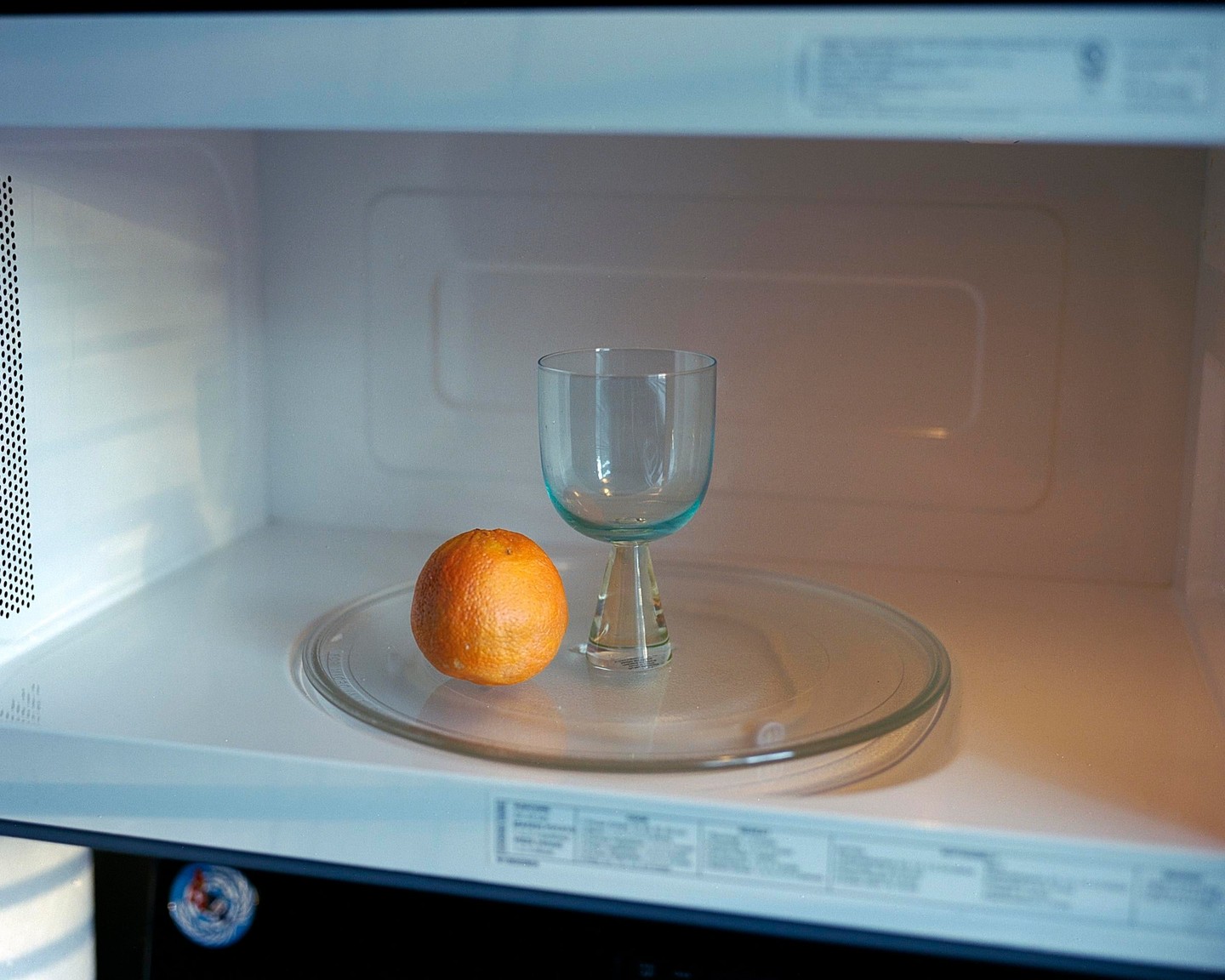 An orange and a glass goblet sit on a glass turntable inside a microwave oven.
