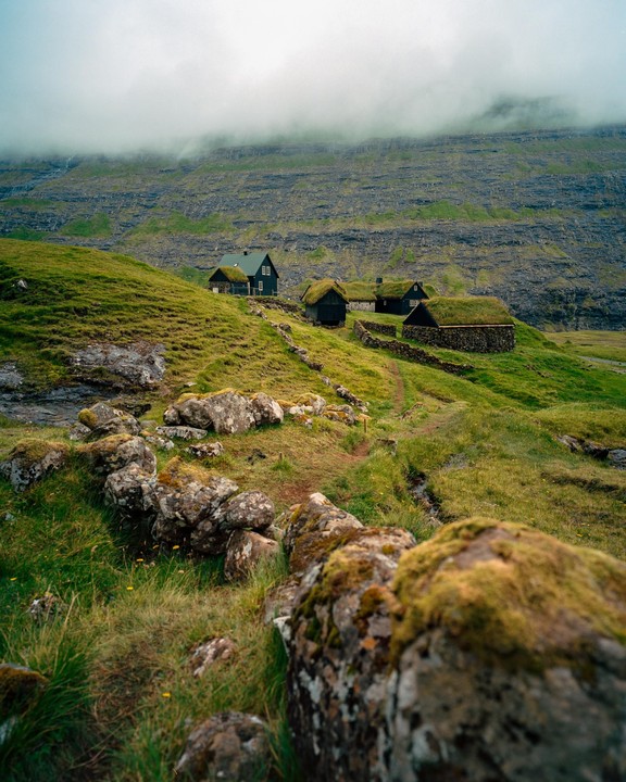 Several traditional houses with grass roofs are nestled into a green hillside under a cloudy sky.