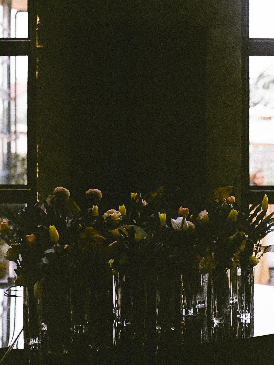 A row of flower arrangements in glass vases sits in front of a window.