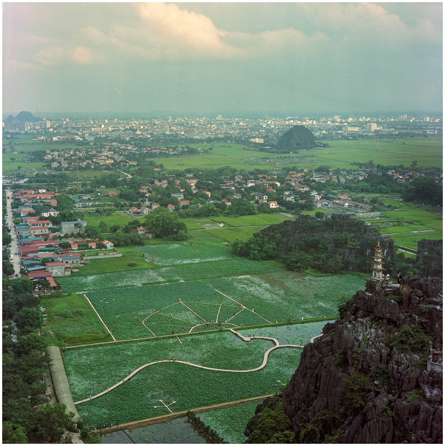 A high-angle landscape shows a city and village surrounded by green fields, with a pagoda on a rocky outcrop in the foreground.