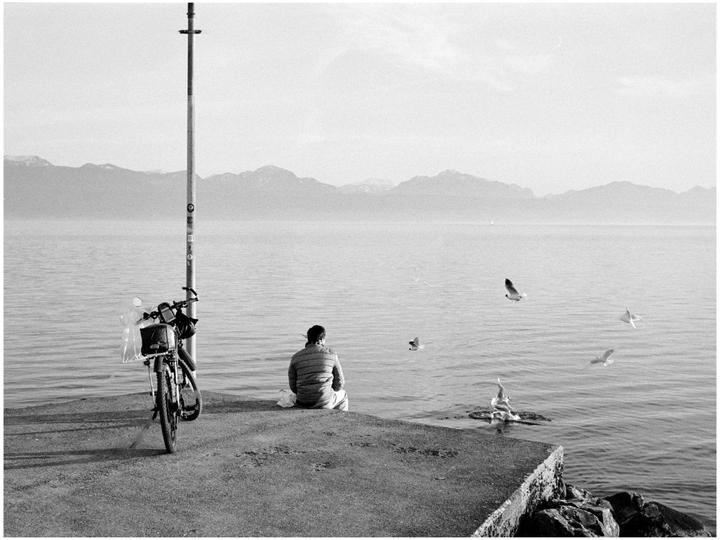 A man sits on a concrete pier next to a bicycle, looking out at a lake with mountains in the distance and several birds flying over the water.