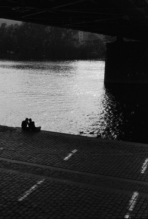 Two people sit on a cobblestone riverbank under a bridge, with sunlight reflecting on the water.