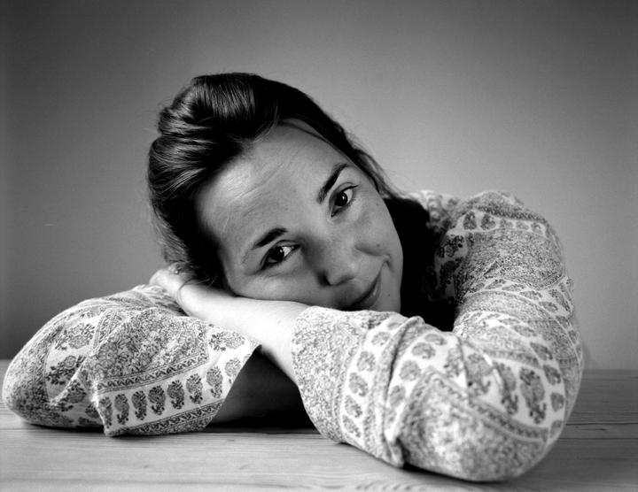 A woman with dark hair rests her head on her arms on a wooden surface, looking towards the viewer with a slight smile.
