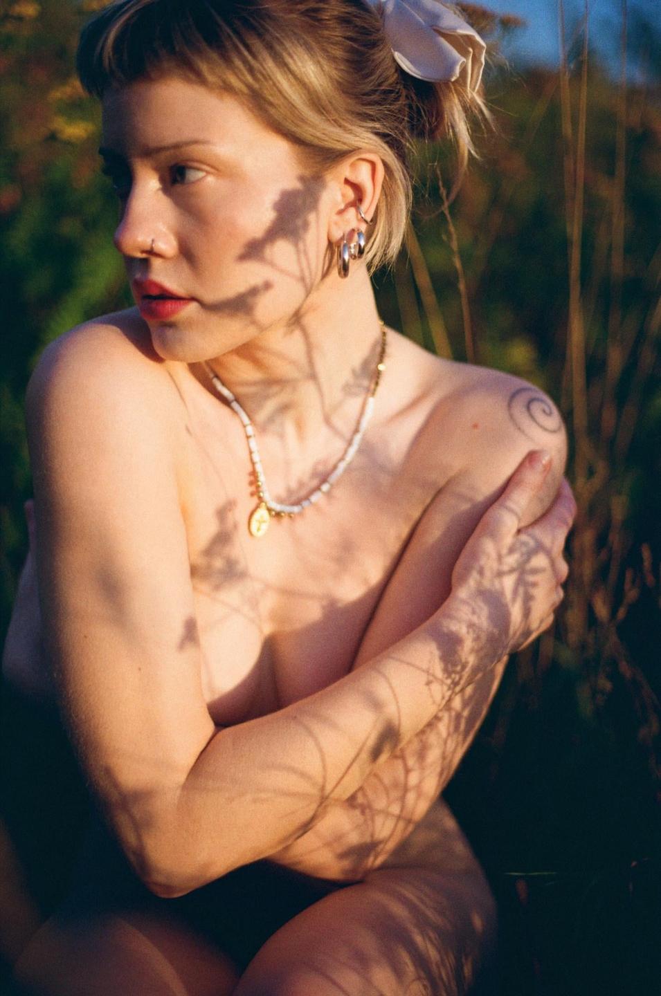 A woman with a nose ring and necklace is sitting in tall grass with shadows of foliage cast upon her body.