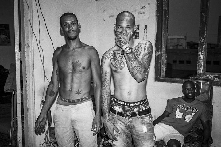 Two men stand shirtless with tattoos while another man sits in a room with a window.