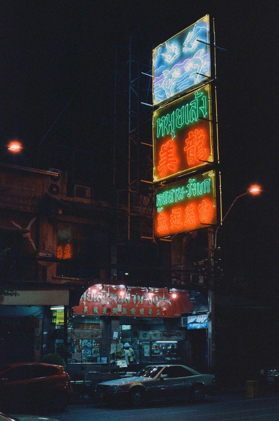 Neon signs illuminate a street at night, with cars parked along the curb and people visible inside a building.