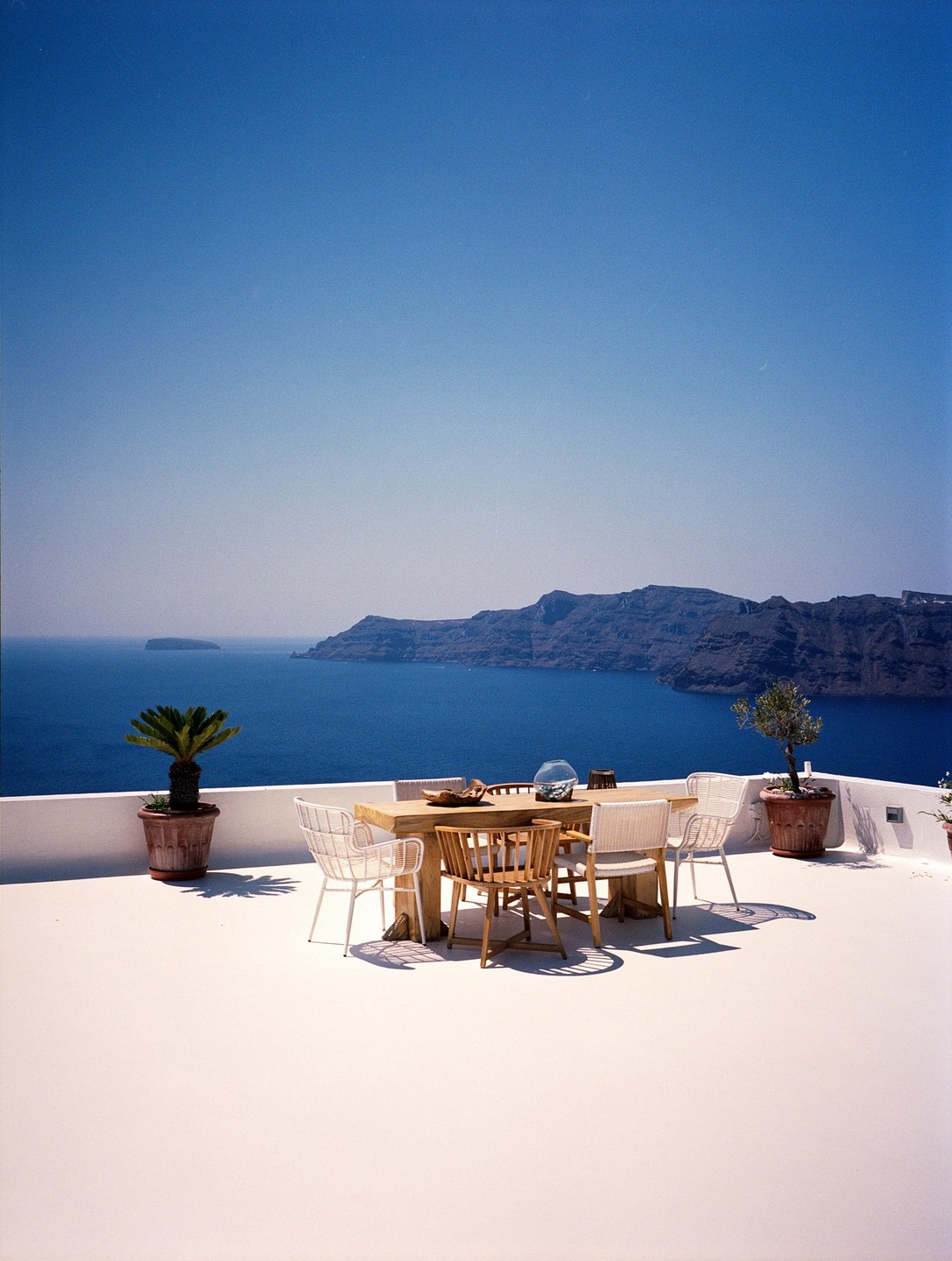 A dining table and chairs sit on a white patio overlooking a vast blue sea and distant mountains.