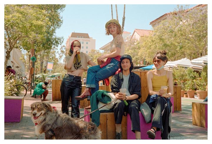 A group of young adults and a dog are gathered on a sunny street with buildings and trees in the background.