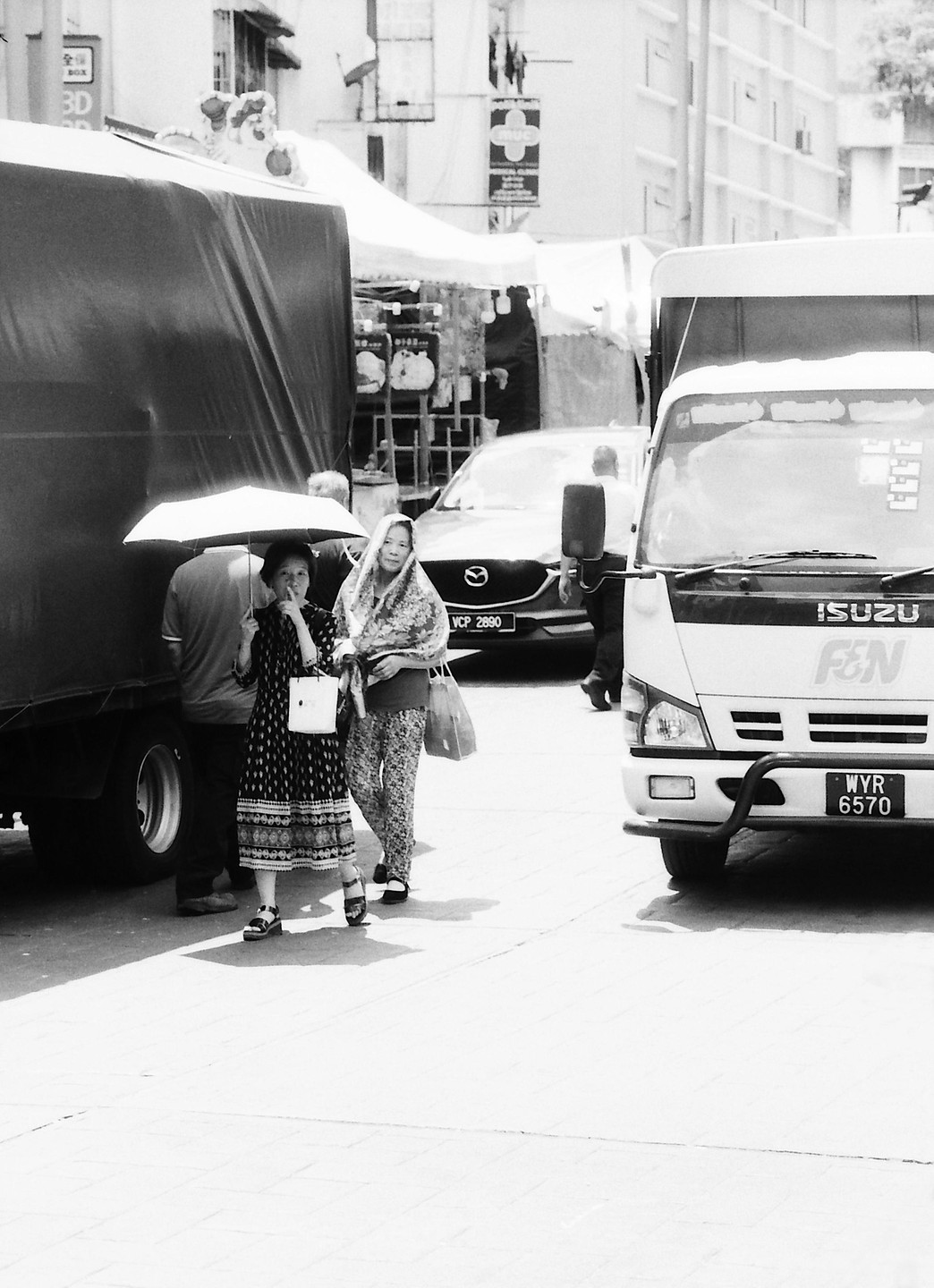 Two women walk on a sunny street, one holding an umbrella, with trucks and cars around them.
