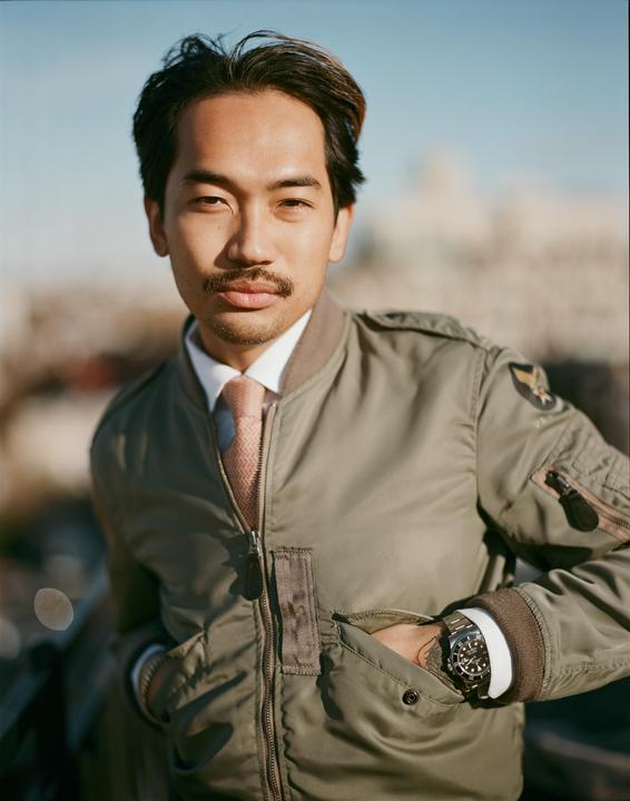 A man wearing a bomber jacket and tie looks directly at the viewer.
