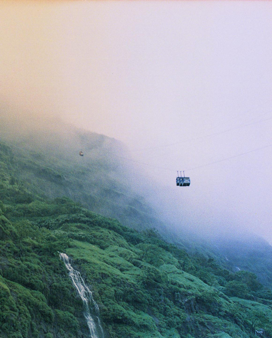 A cable car travels above a lush green mountain with a waterfall, partially obscured by fog.