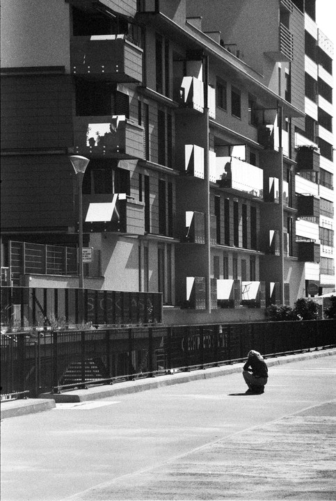 A person crouches on a street next to a fence with apartment buildings in the background.