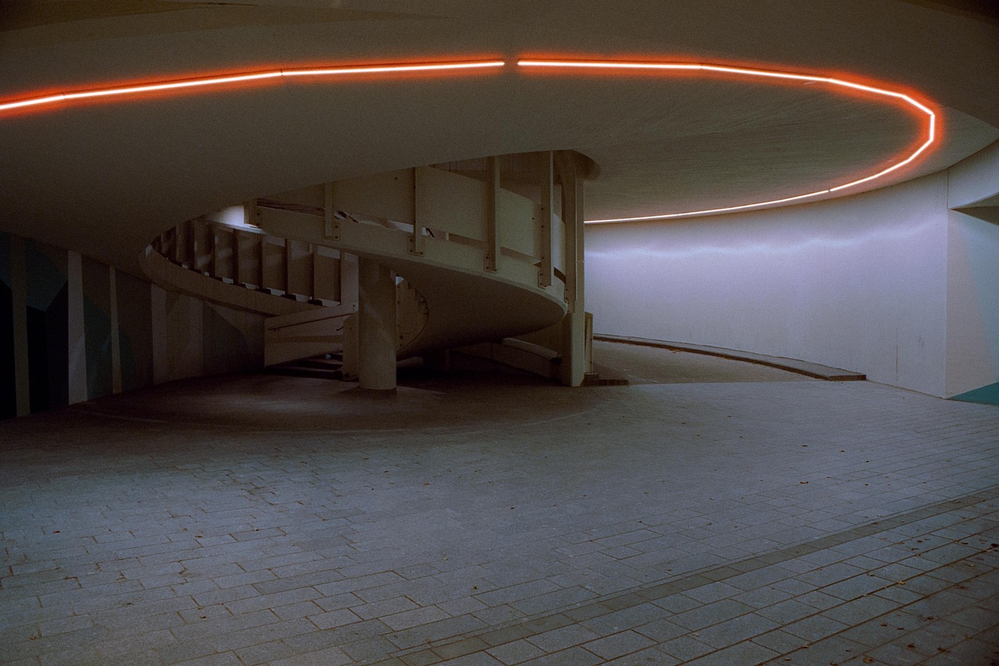 A spiral staircase is illuminated by a neon orange light strip on the ceiling of an underground passage.