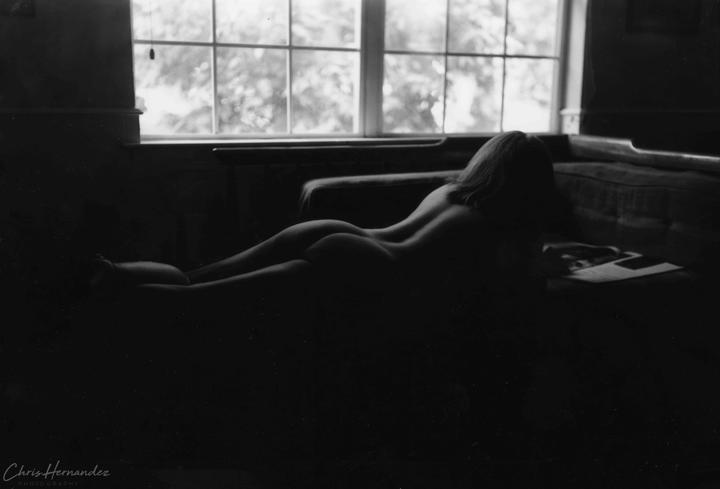 A nude woman lies on a couch with her back to the camera, reading a magazine by a window.