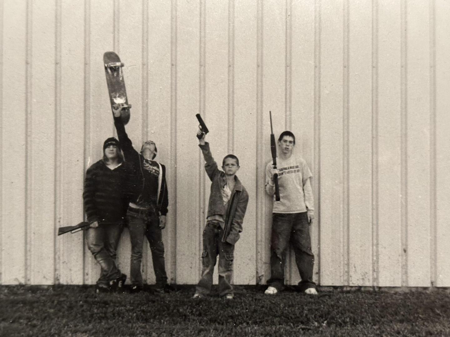 Four boys stand against a corrugated metal wall, two holding rifles, one holding a pistol, and one holding a skateboard.
