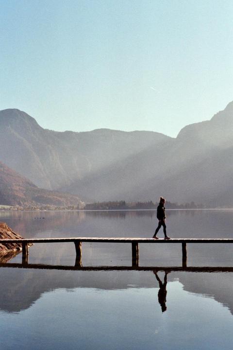 A woman walks across a wooden pier over a lake with mountains in the background.