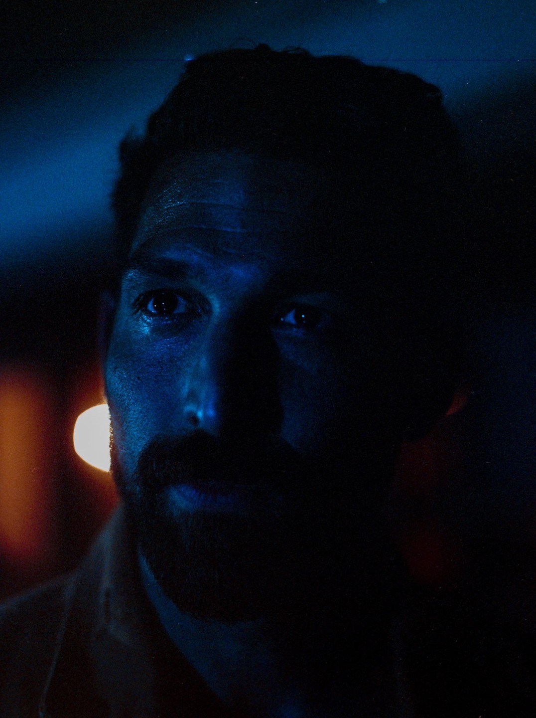 A man with a beard is illuminated by blue light on one side of his face, with a bright orange light source in the background.