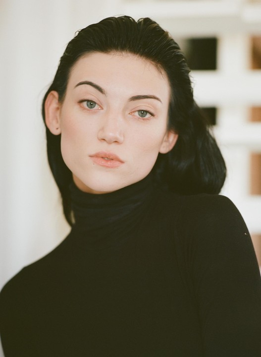 A woman with dark hair and a black turtleneck looks directly at the viewer.